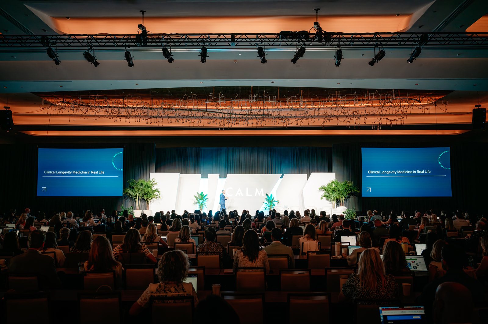 The general session from the back of the room: a full house facing the lit CALM set and two side screens under the lighting truss