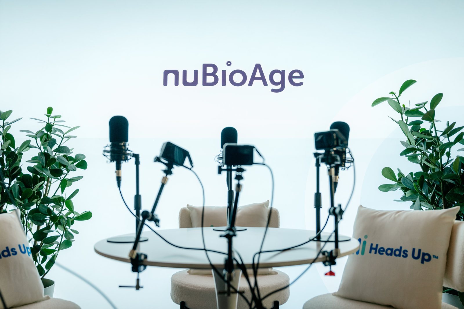 The nuBioAge podcast set: microphones on stands around a round table, white chairs and plants under the nuBioAge mark