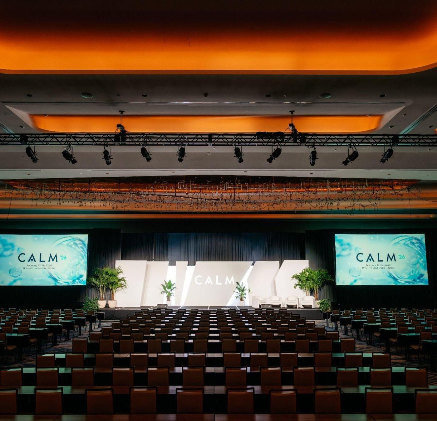 A hotel ballroom set for the CALM 2026 general session: rows of chairs, a white angular stage set, two screens and a lighting truss