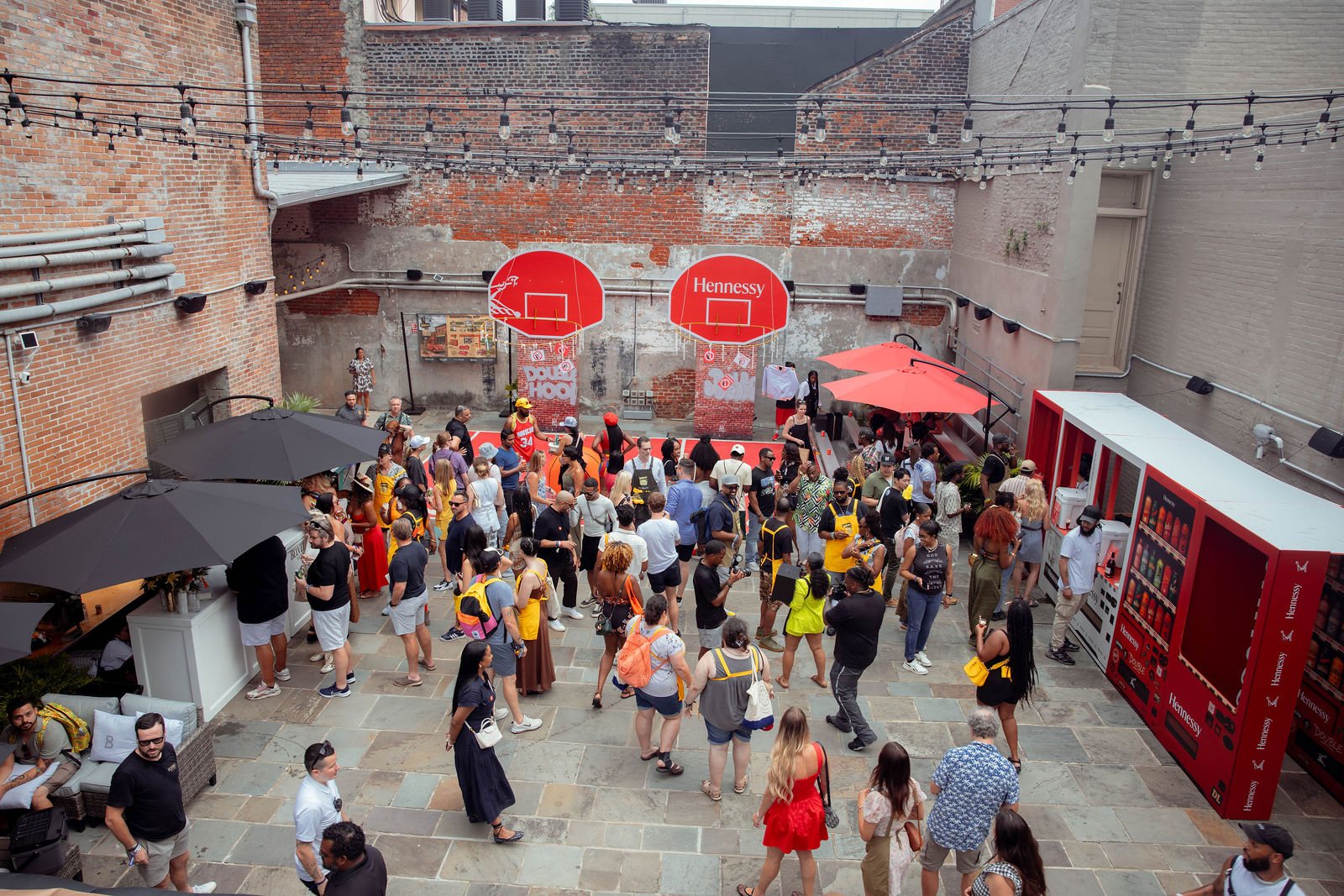 The New Orleans Athletic Club courtyard from above, dressed and full: the double-hoop wall at the far end, red umbrellas, the vending-machine bar at right and the crowd on the court