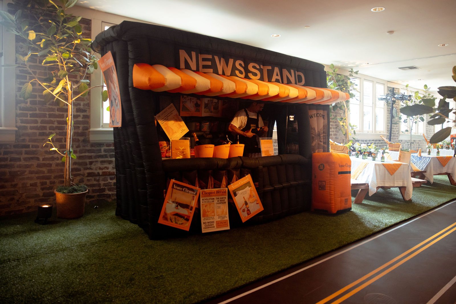 The inflatable newsstand bar, whole: the striped awning, the NEWSSTAND sign, menu boards and orange buckets, on turf with a road-stripe runner