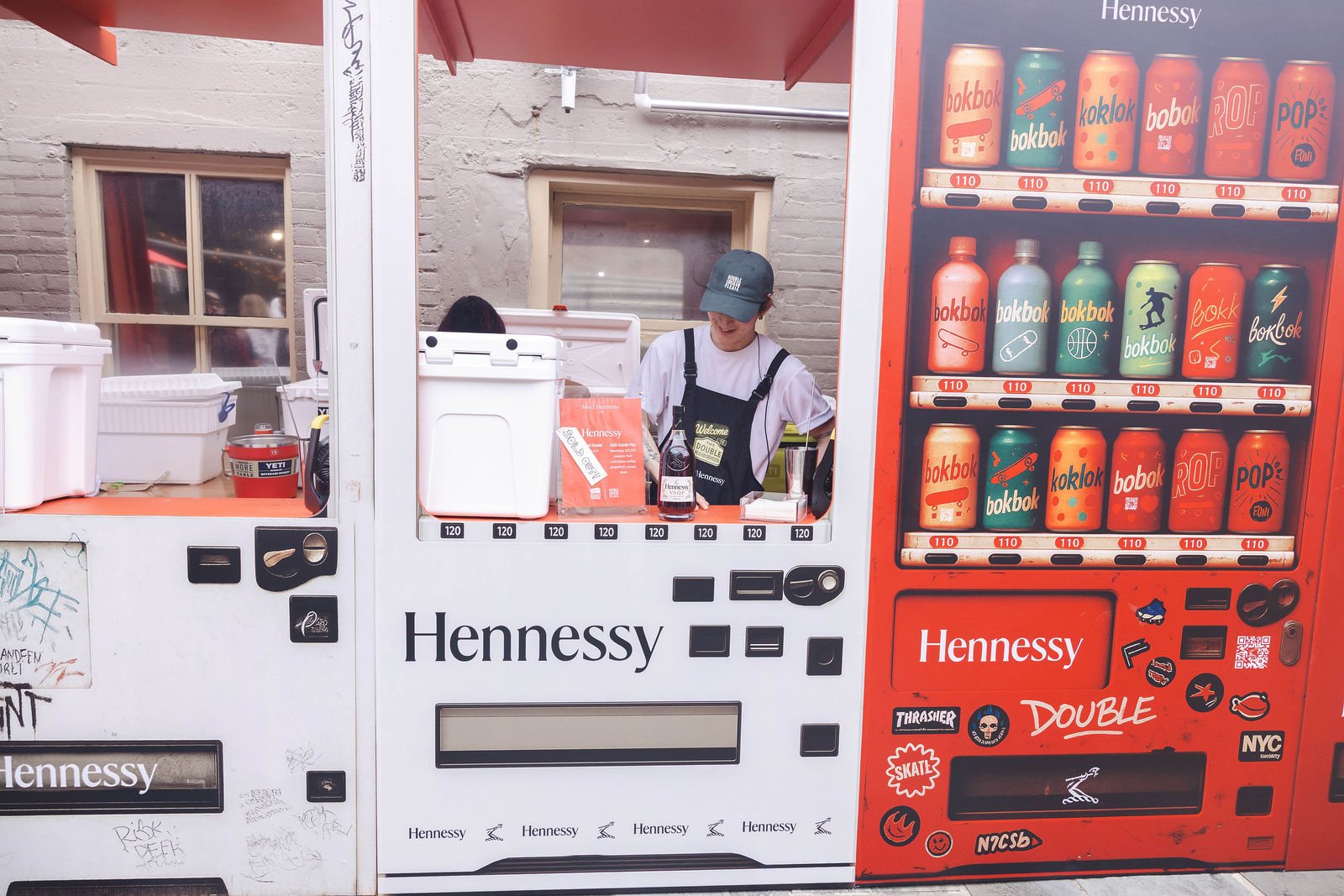 Both vending-machine bars side by side, the white Hennessy unit and the red one, with a bartender serving through the window