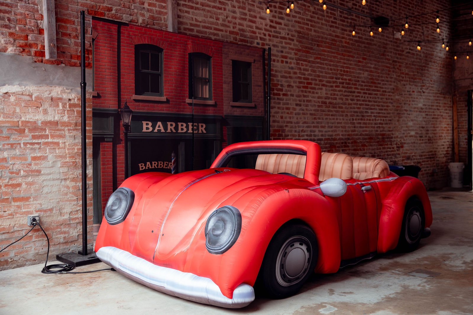 A giant red inflatable convertible parked in front of a painted barber-shop storefront, in a brick room