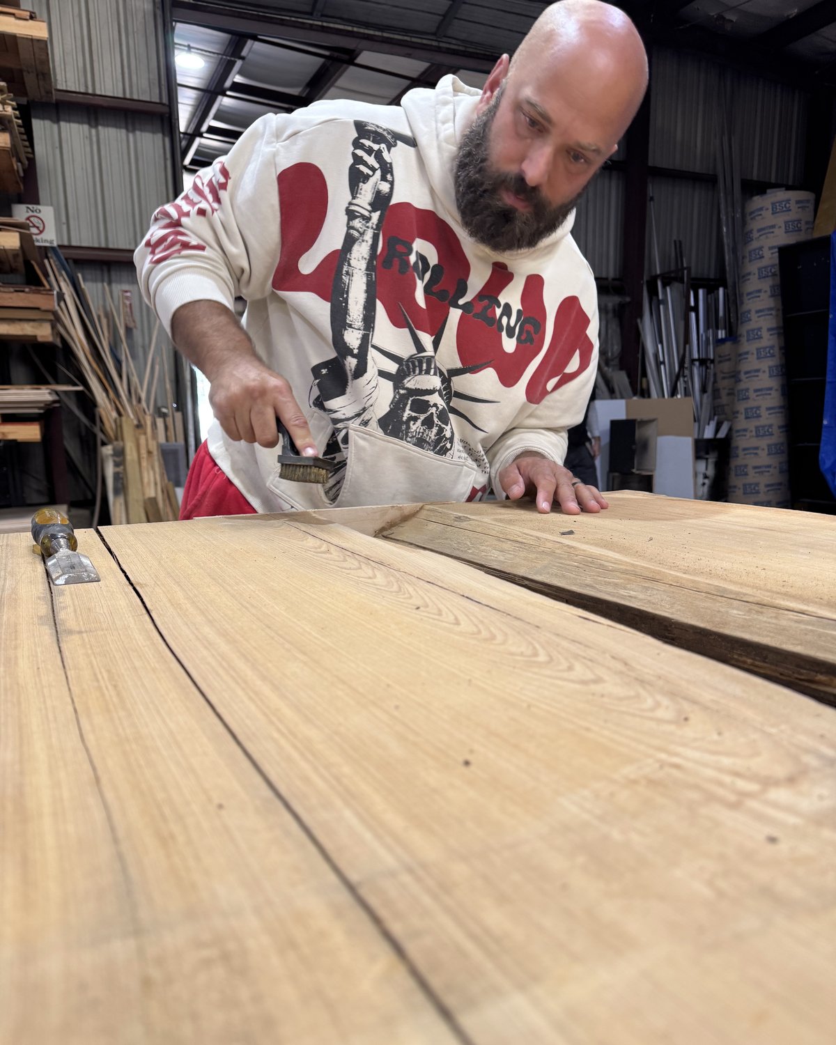 Tim McTee in the shop, brushing down a wood slab on the bench, a chisel beside it, lumber racks behind