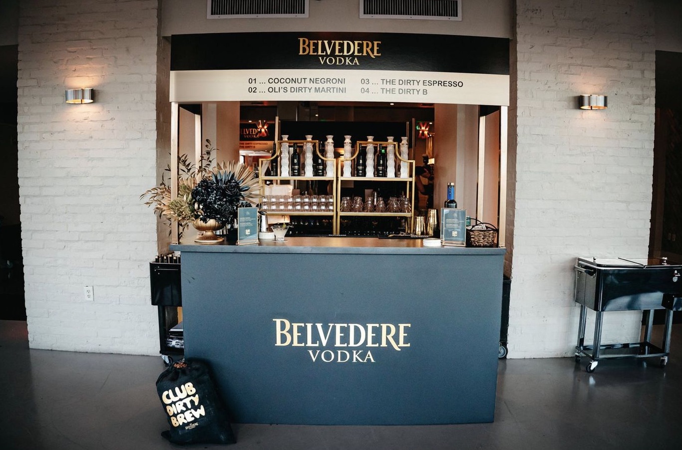 The second custom bar in a white-brick alcove: Belvedere Vodka on the front, a menu of four drinks above, brass rack behind