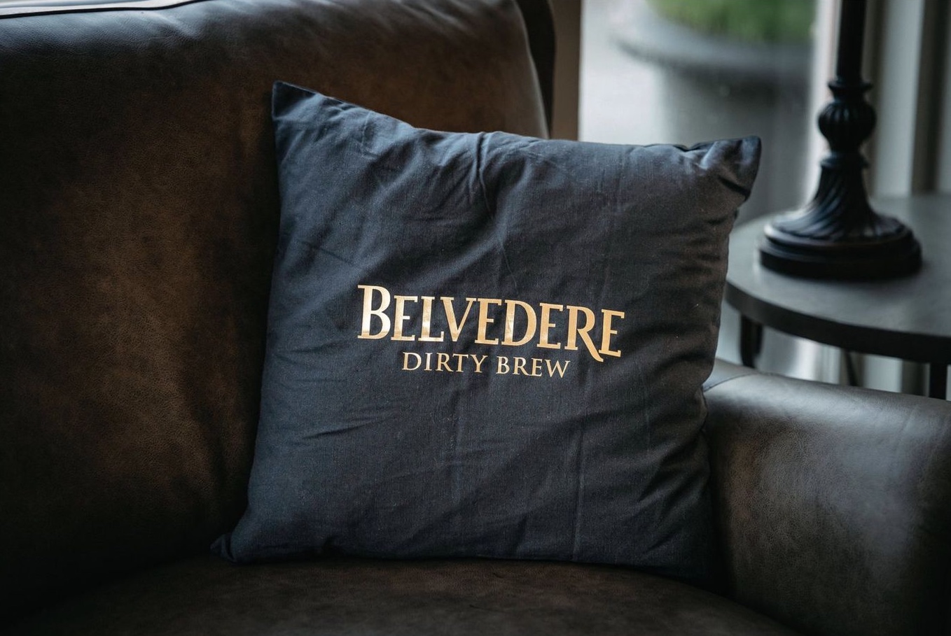 A black pillow printed BELVEDERE DIRTY BREW in gold on a leather armchair